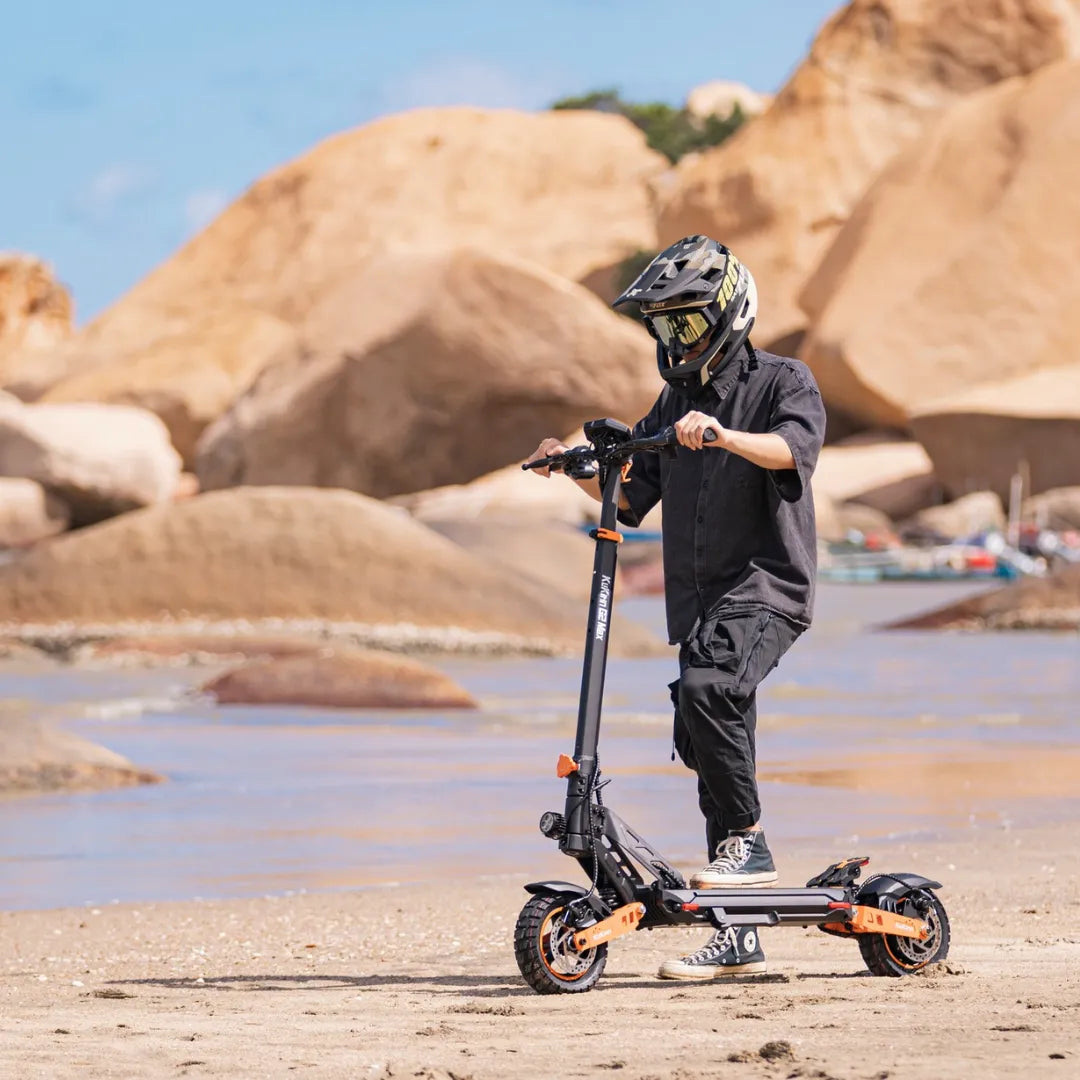 Oseba se vozi z električnim skirojem Kukirin G2 Max na peščeni plaži s skalnatimi formacijami.