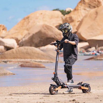 Oseba se vozi z električnim skirojem Kukirin G2 Max na peščeni plaži s skalnatimi formacijami.