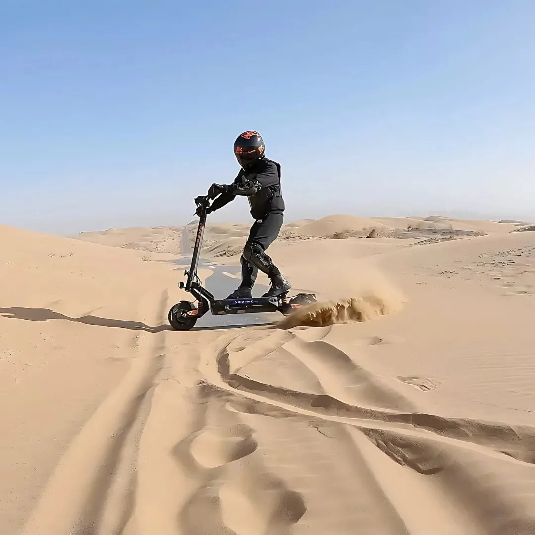 Izdelek na sliki je električni skiro, model Kukirin G2 Master. Vidimo voznika, oblečenega v zaščitno opremo, ki se vozi po peščeni pokrajini. Skuter se premika hitro, kar povzroča prah in pesek, ki se dviga okoli njega. V ozadju so peščene sipine in modro nebo, kar ustvarja dinamično in pustolovsko vzdušje.