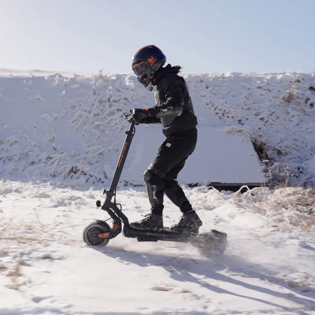 Izdelek na sliki je električni skiro znamke Kukirin, model G2 Master. Na sliki je vidna oseba, ki vozi skiro po zasneženi površini, oblečena v zaščitno opremo, vključno s čelado in zaščitnimi blazinicami. Okolica je prekrita s snegom, kar kaže na zimske razmere.