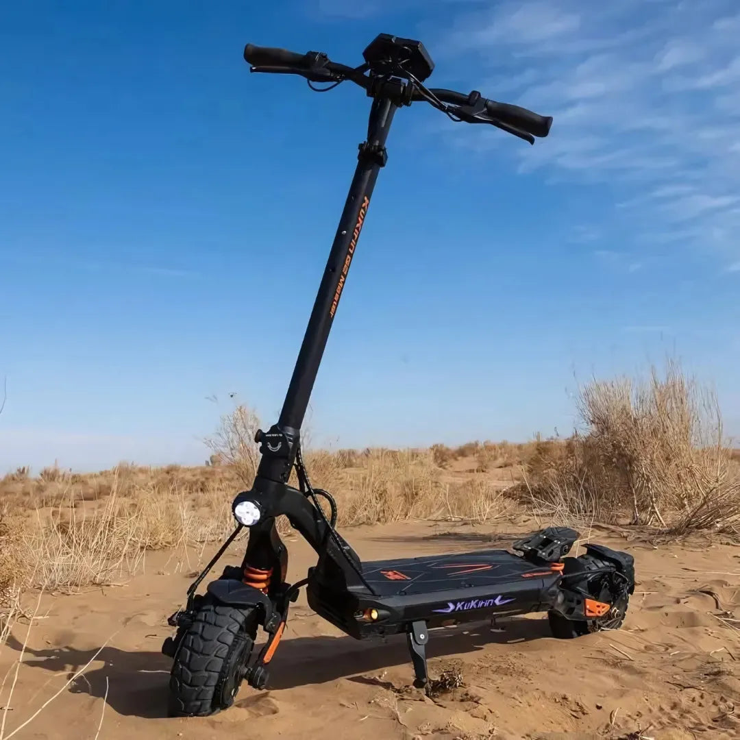 Na sliki je električni skiro, model Kukirin G2 Master. Izdelek ima robustno zasnovo z širokimi pnevmatikami, kar omogoča vožnjo po različnih terenih. Vidna je tudi krmilna enota z ročaji in svetilka na sprednjem delu skiroja. Ozadje prikazuje peščeno okolje z nekaj rastlinjem, kar nakazuje, da je skiro primeren za vožnjo tudi v naravi.