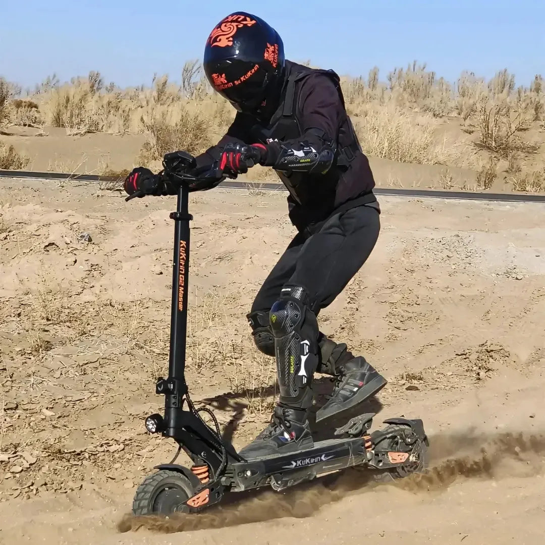 Na sliki je prikazan električni skiro, model Kukirin G2 Master. Oseba na skiroju nosi zaščitno opremo, vključno s čelado in zaščitnimi koleni, ter se vozi po pesku. Skira ima robustno konstrukcijo in široke pnevmatike, kar omogoča vožnjo po različnih terenih. V ozadju so vidne suhe rastline in proga.