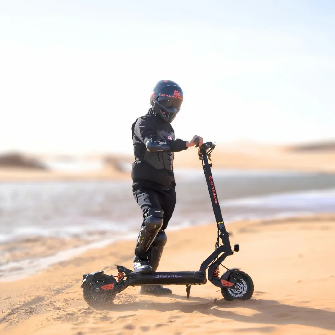 Na sliki je električni skiro, model Kukirin G2 Master. Vidimo otroka, oblečenega v zaščitno opremo, ki se vozi po peščeni površini ob vodi. Skrbno je zaščiten z čelado in zaščitnimi blazinicami na kolenih, kar kaže na varno uporabo skiroja v naravi.