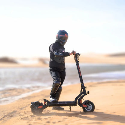 Na sliki je električni skiro, model Kukirin G2 Master. Vidimo otroka, oblečenega v zaščitno opremo, ki se vozi po peščeni površini ob vodi. Skrbno je zaščiten z čelado in zaščitnimi blazinicami na kolenih, kar kaže na varno uporabo skiroja v naravi.