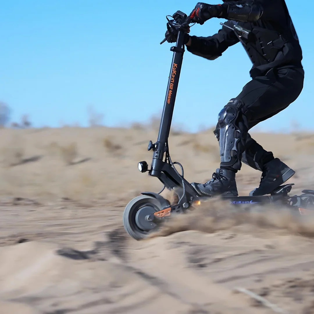 Na sliki je prikazan električni skiro znamke Kukirin, model G2 Master. Uporabnik vozi skiro po peščenem terenu, kar kaže na njegovo sposobnost vožnje po različnih površinah. Vidni so tudi zaščitni elementi, kot so kolenčniki in čelada, kar nakazuje na varno uporabo pri adrenalinskih aktivnostih.