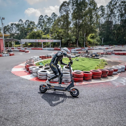 Na sliki je prikazan električni skiro, model Kukirin G4. Na skiroju je oseba v zaščitni opremi, ki se vozi po dirkališču ob robu, obdanim s pnevmatikami. Ozadje prikazuje zeleno območje in druge elemente dirkališča, kar daje občutek dinamike in hitrosti.