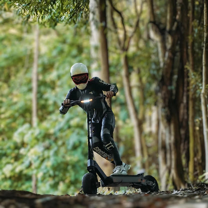 Na sliki je prikazan električni skiro, model Kukirin G4. Uporabnik, oblečen v črno zaščitno obleko in čelado, vozi skiro po gozdni poti. Okolica je zelena in naravna, kar nakazuje na primernost skiroja za vožnjo v naravi. Na skiroju so vidne tudi LED luči, kar povečuje njegovo vidnost.