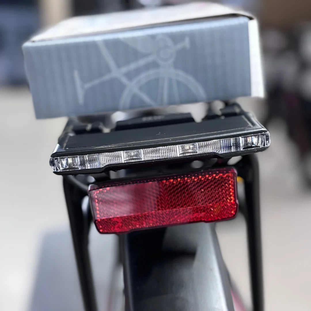 Izdelek na sliki je električno kolo znamke Malaguti, model Bolonina. Na sliki je vidna zadnja luč in odsevnik, ki sta nameščena na okvirju kolesa. V ozadju je tudi škatla, ki verjetno vsebuje dodatno opremo ali navodila. Luči in odsevnik sta pomembna za varnost vožnje v temi.