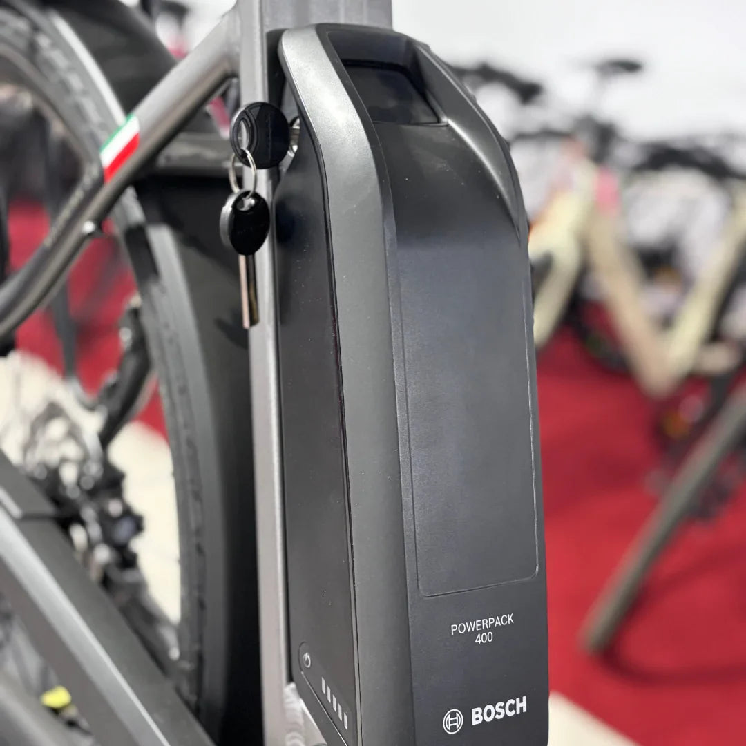 Izdelek je električno kolo znamke Malaguti, model Bolonina. Na sliki je prikazana baterija Bosch Powerpack 400, ki je nameščena na okvirju kolesa. Baterija ima ključek in indikator napolnjenosti, kar nakazuje na njeno funkcionalnost in enostavno uporabo. V ozadju so vidna tudi druga kolesa, kar daje občutek razstave.