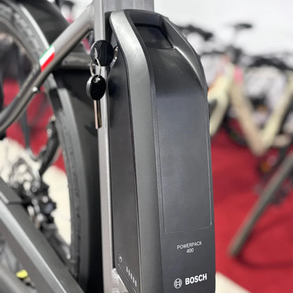 Izdelek je električno kolo znamke Malaguti, model Bolonina. Na sliki je prikazana baterija Bosch Powerpack 400, ki je nameščena na okvirju kolesa. Baterija ima ključek in indikator napolnjenosti, kar nakazuje na njeno funkcionalnost in enostavno uporabo. V ozadju so vidna tudi druga kolesa, kar daje občutek razstave.