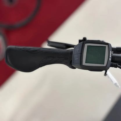 Na sliki je električno kolo znamke Malaguti, model Bolonina. Vidna je ročica krmila s črnim oprijemom in zaslon, ki prikazuje informacije o delovanju kolesa. Zaslon ima gumbe za prilagajanje, kar nakazuje na funkcionalnosti, kot so izbira hitrosti ali način vožnje. Ozadje slike je nejasno, vendar se zdi, da je prostor urejen in primeren za predstavitev kolesa.