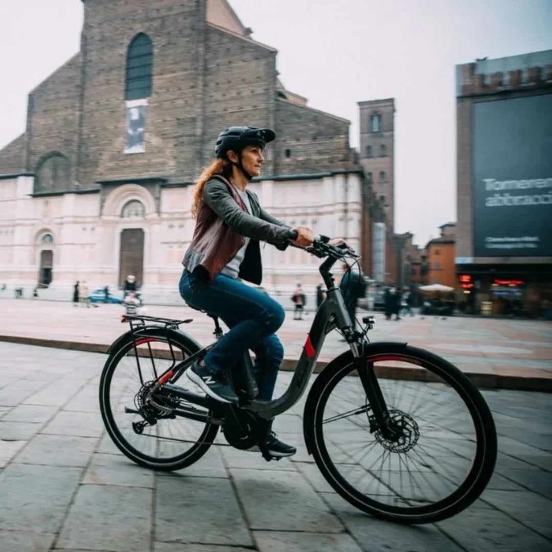 Na sliki je električno kolo znamke Malaguti, model Bolonina. Prikazuje žensko, ki vozi kolo po mestnem trgu, obdanem z arhitekturnimi znamenitostmi. Kolo ima sodoben dizajn in je opremljeno z električnim motorjem ter udobnim sedežem, kar omogoča prijetno vožnjo. Oblečena je v udobna oblačila in čelado, kar kaže na zavedanje o varnosti med vožnjo.