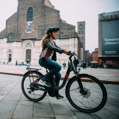 Na sliki je električno kolo znamke Malaguti, model Bolonina. Prikazuje žensko, ki vozi kolo po mestnem trgu, obdanem z arhitekturnimi znamenitostmi. Kolo ima sodoben dizajn in je opremljeno z električnim motorjem ter udobnim sedežem, kar omogoča prijetno vožnjo. Oblečena je v udobna oblačila in čelado, kar kaže na zavedanje o varnosti med vožnjo.