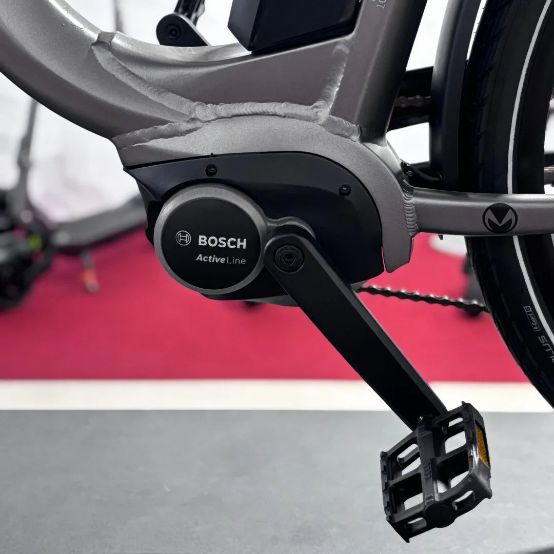 Na sliki je električno kolo znamke Malaguti, model Bolonina. Vidna je motorna enota Bosch Active Line, ki se nahaja na spodnjem delu okvirja, skupaj s pedalom. Kolo ima sodobno obliko in robustno konstrukcijo, kar nakazuje na kakovost in zmogljivost. Ozadje je nejasno, kar poudari izdelek.