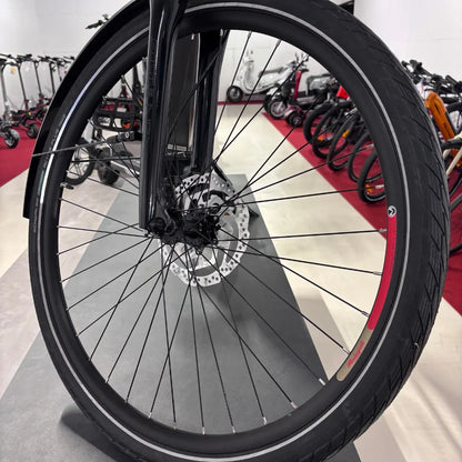 Izdelek na sliki je električno kolo znamke Malaguti, model Bolonina. Na sliki je prikazan sprednji kolo, ki vključuje disk zavore in pnevmatik z značilnim profilom. V ozadju so vidni tudi drugi električni skiroji in kolesa, kar nakazuje, da gre za prodajno mesto ali razstavo.