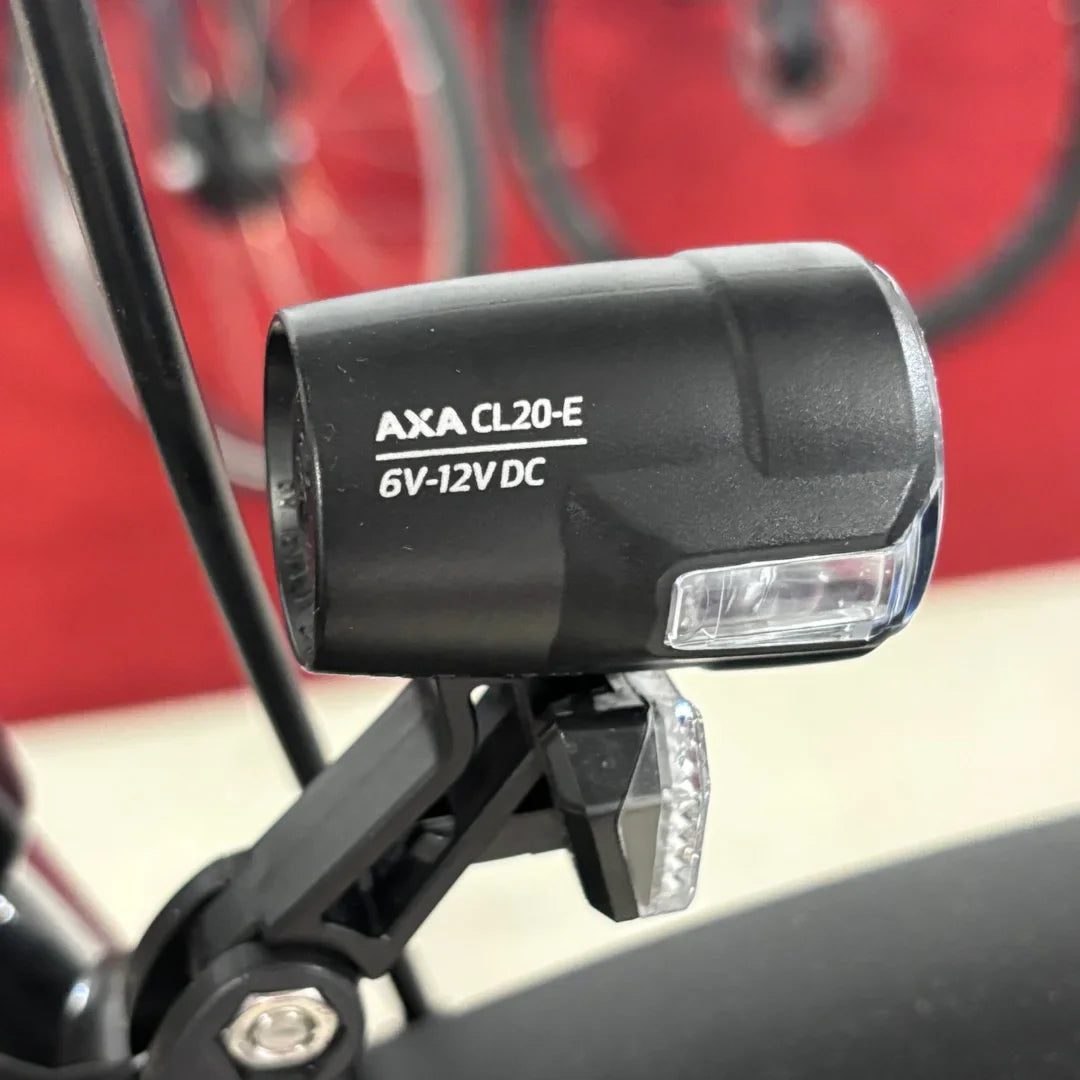 Na sliki je prikazana luč za električno kolo znamke AXA, model CL20-E, ki deluje na napetosti 6V-12V DC. Luč je nameščena na okvir kolesa in je zasnovana za izboljšanje vidnosti med vožnjo. V ozadju so delno vidni kolesi, kar nakazuje, da gre za električno kolo.
