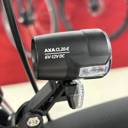 Na sliki je prikazana luč za električno kolo znamke AXA, model CL20-E, ki deluje na napetosti 6V-12V DC. Luč je nameščena na okvir kolesa in je zasnovana za izboljšanje vidnosti med vožnjo. V ozadju so delno vidni kolesi, kar nakazuje, da gre za električno kolo.
