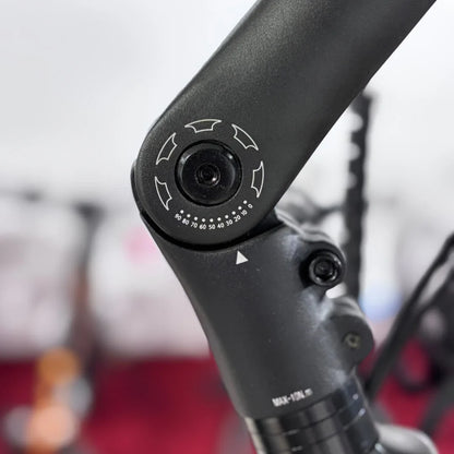 Na sliki je električno kolo znamke Malaguti, model Bolonina. Vidna je podrobnost na okvirju, kjer se nahaja nastavitveni gumb, ki omogoča prilagajanje različnih funkcij kolesa. Gumb ima označene vrednosti in smeri, kar kaže na možnost prilagoditve nastavitve. Ozadje slike je zamejeno, kar daje poudarek na kolesu.