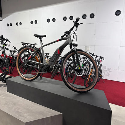 Na sliki je električno kolo znamke Malaguti, model Carezza TR 4.0 High Step. Kolo je postavljeno na stojalo v trgovini, kar omogoča dober pregled nad njegovim dizajnom. Ima robusten okvir v sivi barvi s poudarki v rdeči barvi, široke pnevmatike in je primerno za vožnjo po različnih terenih. V ozadju so vidna tudi druga kolesa, kar nakazuje na raznoliko izbiro izdelkov v trgovini.