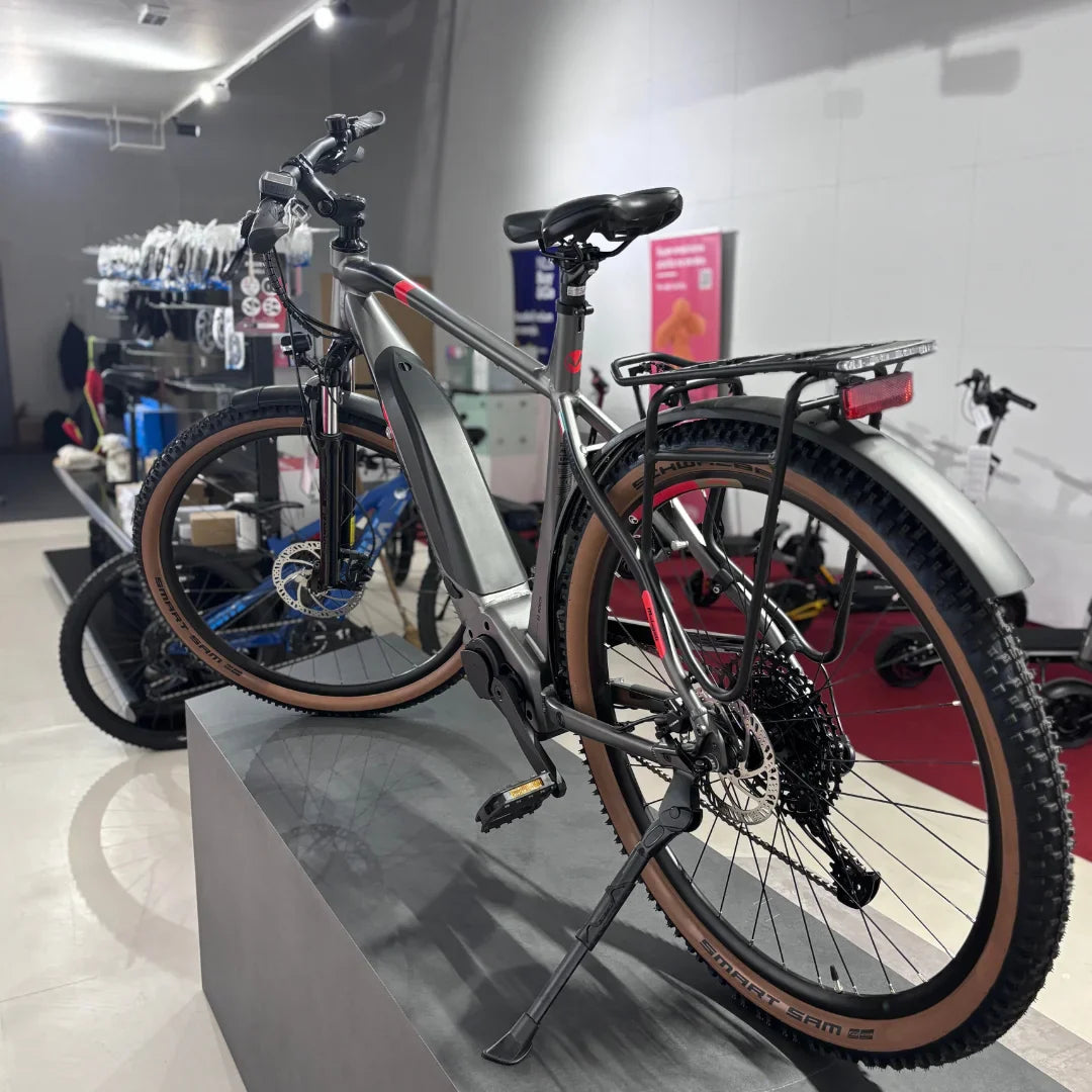 Izdelek na sliki je električno kolo znamke Malaguti, model Carezza TR 4.0 z visokim korakom. Kolo ima moderno zasnovo, opremljeno z električnim motorjem in baterijo, kar omogoča enostavno in udobno vožnjo. Na sliki je vidna tudi zadnja prtljažna mreža ter široki kolesi z gumami, ki so primerne za različne terene. V ozadju so vidni drugi kolesi in oprema, kar nakazuje, da gre za prodajno mesto ali servis.