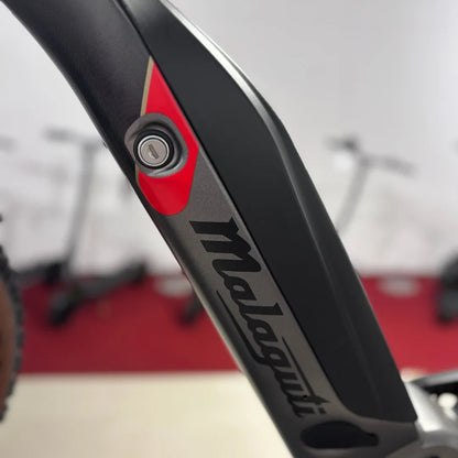 Izdelek na sliki je električno kolo, znamke Malaguti, model Carezza TR 4.0 High Step. Na sliki je vidna del okvirja, kjer je nameščen ključavnica za baterijo, kar nakazuje na električni sistem kolesa. Obkroža ga rdeča in črna barva, kar daje kolesu sodoben videz. V ozadju so vidni tudi drugi kolesi.