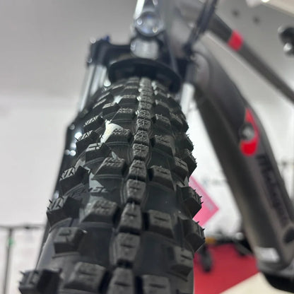 Izdelek je električno kolo, znamke Malaguti, model Carezza TR 4.0 High Step. Na sliki je viden profil pnevmatike, ki ima izrazit terenski profil, kar nakazuje, da je kolo primerno za vožnjo po različnih površinah. V ozadju se vidi del okvirja kolesa, kar dodatno potrjuje športni videz in robustnost izdelka.