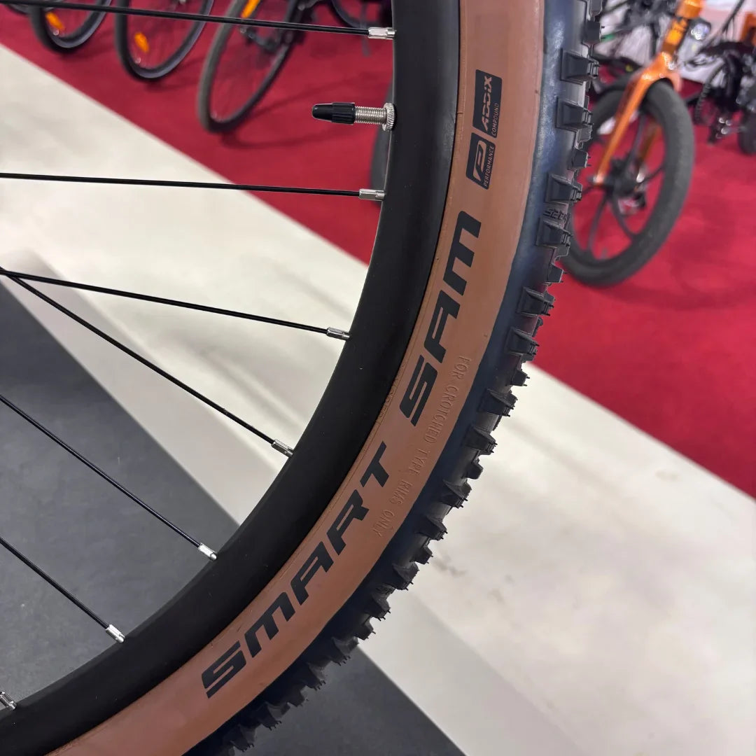 Izdelek na sliki je električno kolo, znamke Malaguti, model Carezza TR 4.0 High Step. Na sliki je del kolesa, natančneje kolo s pnevmatiko, ki nosi oznako "SMART SAM". Pnevmatika ima profil, primeren za različne terene, kar nakazuje na vsestranskost kolesa. V ozadju so vidna še druga kolesa, kar daje občutek o razstavi ali trgovini.