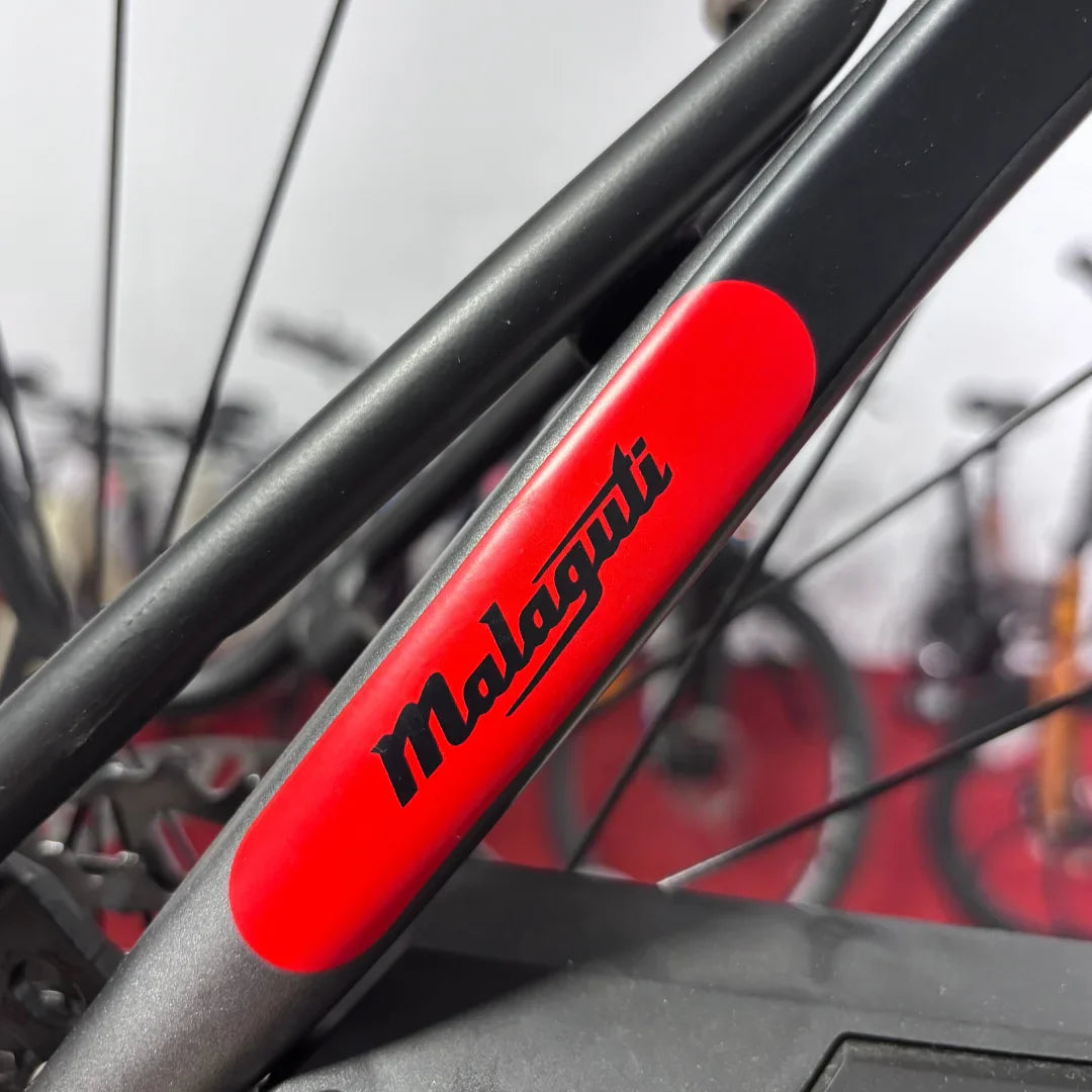 Izdelek na sliki je električno kolo znamke Malaguti, model Carezza TR 4.0 High Step. Na sliki je vidna podrobnost okvirja, kjer je jasno razvidno ime znamke Malaguti v črni barvi na rdečem ozadju. V ozadju so vidne tudi druge kolesarske komponente, kar nakazuje, da je kolo del razstave ali trgovine.