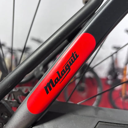 Izdelek na sliki je električno kolo znamke Malaguti, model Carezza TR 4.0 High Step. Na sliki je vidna podrobnost okvirja, kjer je jasno razvidno ime znamke Malaguti v črni barvi na rdečem ozadju. V ozadju so vidne tudi druge kolesarske komponente, kar nakazuje, da je kolo del razstave ali trgovine.