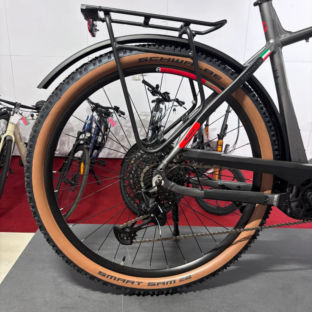 Izdelek na sliki je električno kolo, model Malaguti Carezza TR 4.0 High Step. Na sliki je vidna zadnja stran kolesa z značilnimi širokimi pnevmatikami Schwalbe Smart Sam, ki so primerne za različne terene. Kolo ima tudi prtljažnik in robustno konstrukcijo, kar nakazuje na njegovo uporabnost za daljše vožnje in potovanja. V ozadju so vidna tudi druga kolesa.