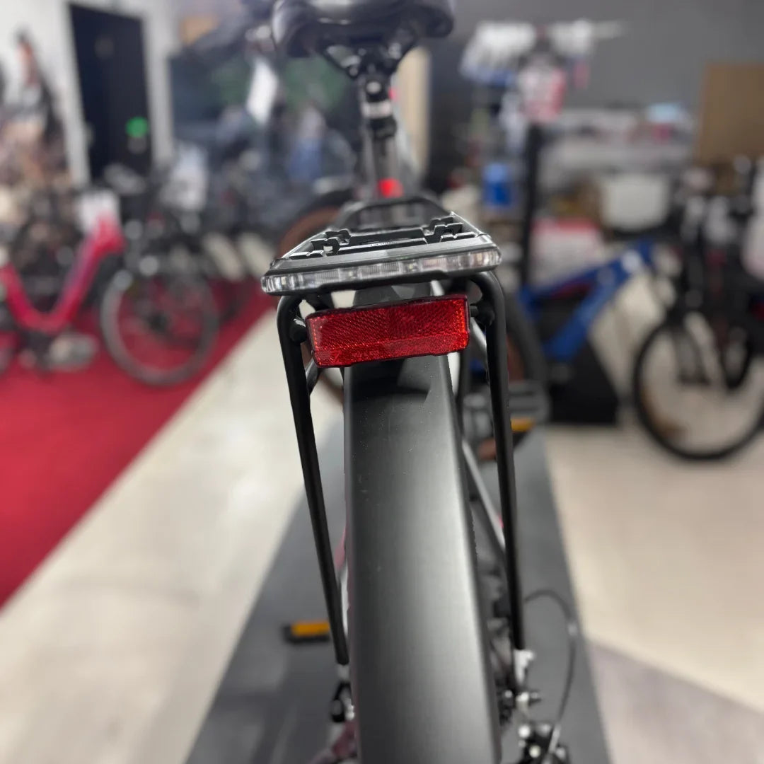 Izdelek na sliki je električno kolo znamke Malaguti, model Carezza TR 4.0 High Step. Na sliki je vidna zadnja stran kolesa, kjer je nameščen nosilec za prtljago in rdeča odsevna lučka, kar nakazuje na funkcionalnost in varnost kolesa. V ozadju so vidni tudi drugi kolesi, kar nakazuje, da je slika posneta v trgovini s kolesi.