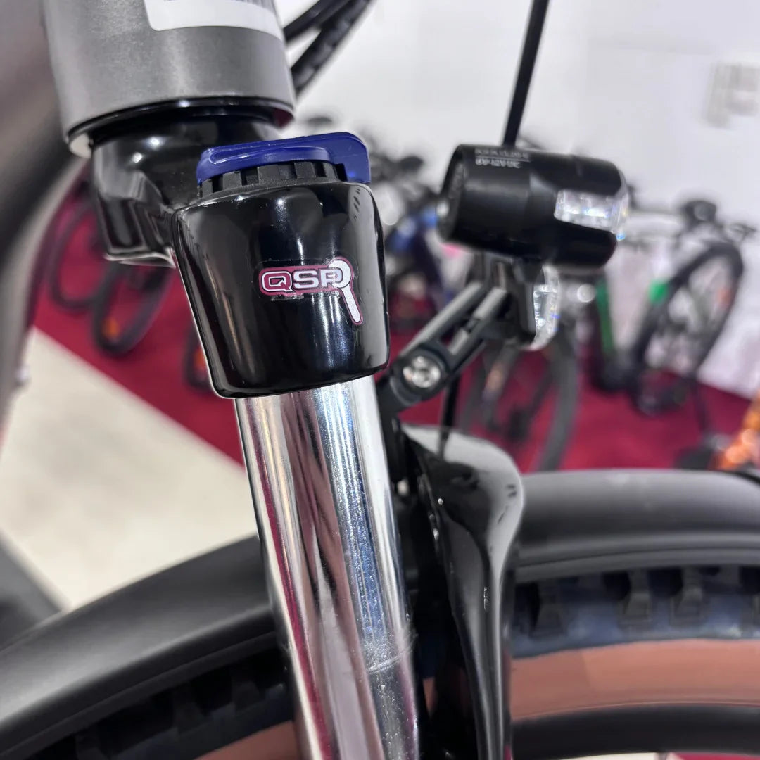 Izdelek na sliki je električno kolo znamke Malaguti, model Carezza TR 4.0 High Step. Na sliki je prikazan del sprednje vilice, ki vključuje sistem za prilagajanje vzmetenja ter del kolesa, kar nakazuje na kakovostno izdelavo in funkcionalnost. V ozadju so vidni tudi drugi kolesi, kar daje občutek o raznolikosti ponudbe.