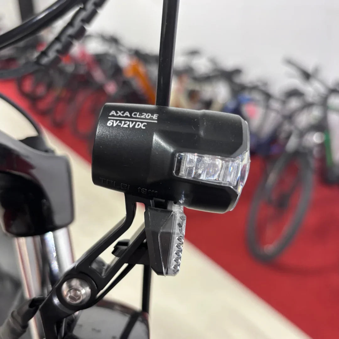 Na sliki je prikazan del električnega kolesa, model Malaguti Carezza TR 4.0 High Step. Vidna je sprednja luč AXA CL20-E, ki je zasnovana za osvetlitev poti med vožnjo. V ozadju so vidna druga kolesa, kar nakazuje, da je slika posneta v trgovini ali razstavišču.