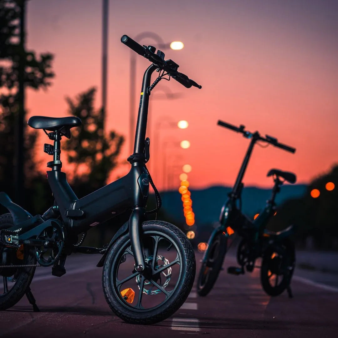 Na sliki je električno kolo, model MS Energy Urbanfold i6. Kolo ima kompakten dizajn s manjšimi kolesi in zložljivo konstrukcijo, kar ga naredi primernega za urbano vožnjo. Na sliki sta vidna dva električna kolesa postavljena na cesti, v ozadju pa je slikovit sončni zahod, kar ustvarja prijetno vzdušje. Kolesi imata temno barvo in sodoben videz, kar poudarja njihovo funkcionalnost in stil.