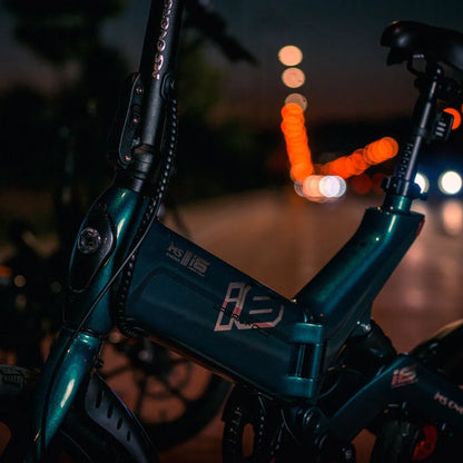 Izdelek na sliki je električno kolo, natančneje model MS Energy Urbanfold i6. Na sliki je vidna podrobnost okvirja kolesa v temno zeleni barvi, ki ima eleganten dizajn. Osvetlitev v ozadju ustvarja prijetno nočno vzdušje, kar nakazuje na možnost vožnje v večernih urah.