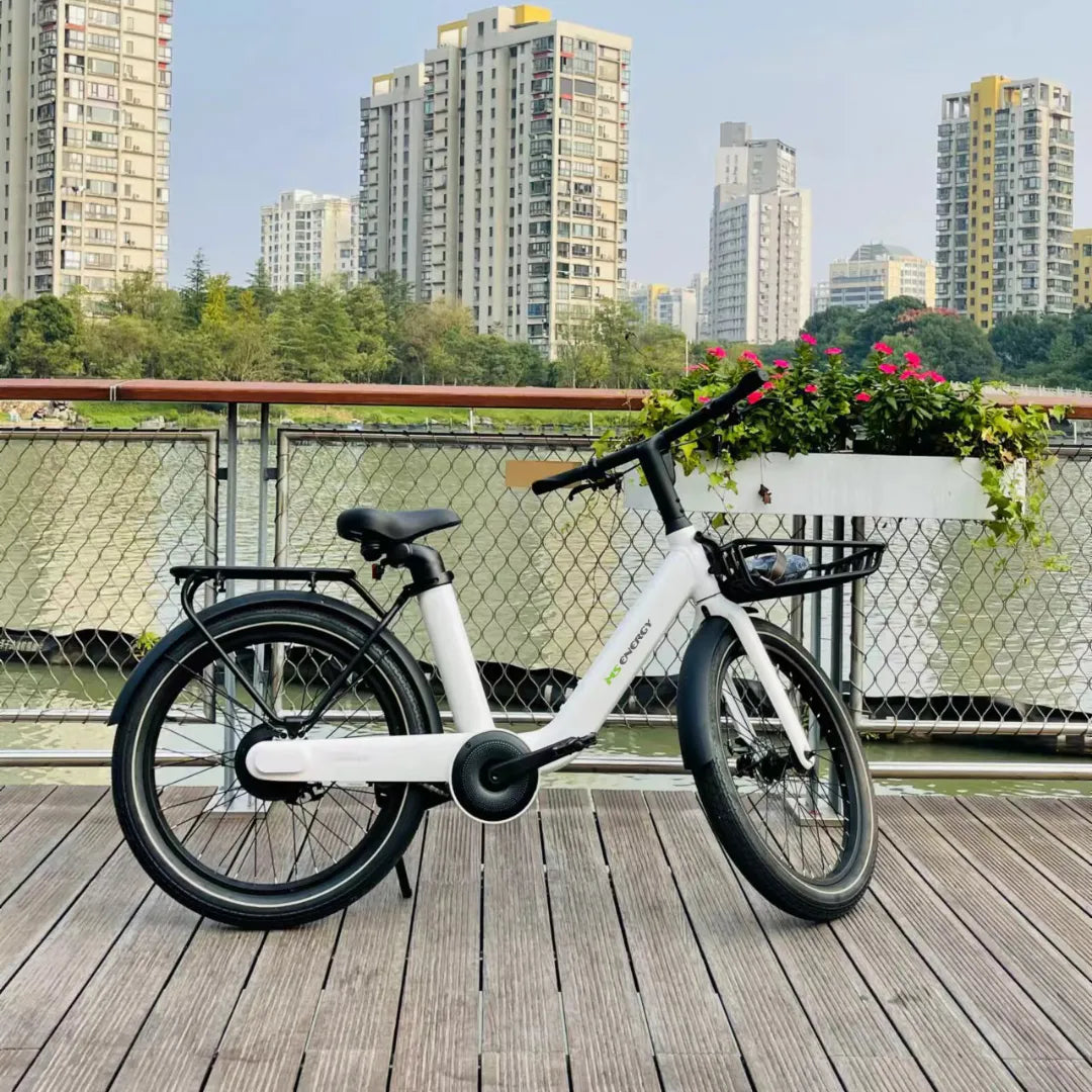 Izdelek na sliki je električno kolo, model MS Energy Cityzen C102. Kolo je belo, ima udoben sedež in široke pnevmatike, kar zagotavlja stabilnost in udobno vožnjo. Na sliki je kolo postavljeno na leseno ploščad ob vodi, v ozadju pa so vidne moderne zgradbe in cvetlična zasaditev.