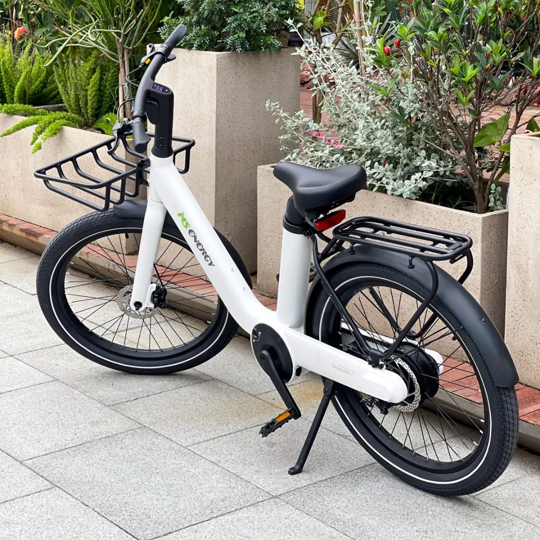 Na sliki je električno kolo znamke MS Energy, model Cityzen C102. Kolo je belo in ima moderno obliko, opremljeno je z dvema prtljažnikoma, sprednjim in zadnjim. V ozadju so vidne zelene rastline, kar daje sliki prijeten naravni občutek. Na krmilu je tudi zaslon, ki verjetno prikazuje informacije o hitrosti ali stanju baterije.