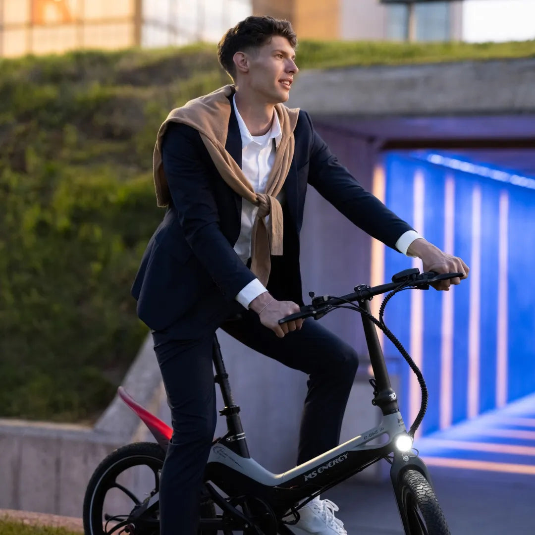 Izdelek na sliki je električno kolo, model MS Energy I20. Na sliki je moški v poslovnem obleku, ki se vozi na kolesu. Kolo ima moderno zasnovo in je opremljeno z lučjo spredaj. V ozadju je arhitekturna struktura, ki ustvarja sodoben ambient.