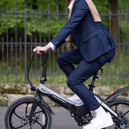 Na sliki je električno kolo znamke MS Energy, model i20. Oseba, oblečena v poslovno obleko, se vozi na kolesu, ki ima kompakten dizajn in manjše kolesa. Vidni so tudi elementi, kot so krmilo in sedež, kar nakazuje na udobno vožnjo. Okolje je zeleno, kar ustvarja prijetno vzdušje za vožnjo.