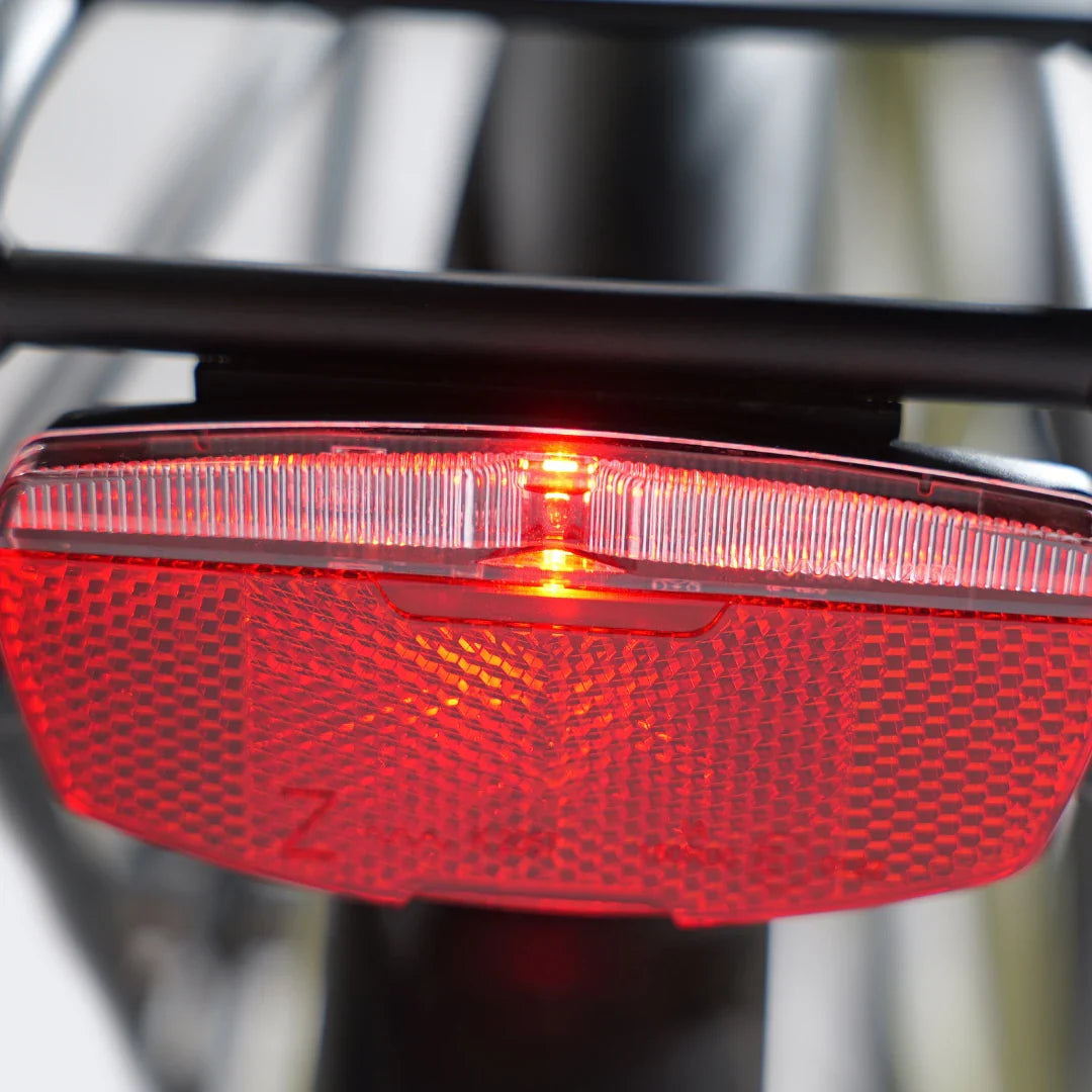 Izdelek na sliki je električno kolo, model MS Energy C300. Na sliki je prikazan zadnji odsevnik in lučka, ki sta del varnostne opreme kolesa. Rdeča lučka je vklopljena, kar povečuje vidnost kolesarja v temi. Odsevnik je zasnovan za dodatno varnost med vožnjo.