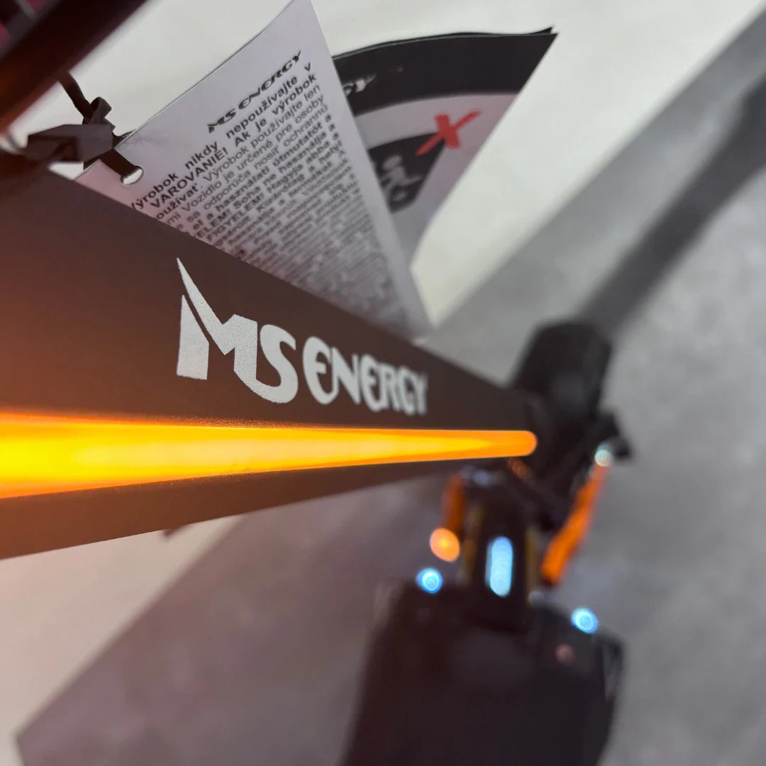 Izdelek na sliki je električni skiro, model MS Energy Flare X Pro. Na sliki je vidna črna konstrukcija skiroja z logotipom znamke MS Energy, ki je opremljena z LED osvetlitvijo v oranžni barvi. V ozadju se vidi tudi navodilo za uporabo, kar nakazuje, da gre za nov izdelek.