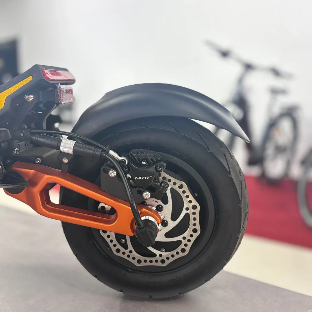 Izdelek na sliki je električni skiro, model MS Energy Flare X Pro. Na sliki je prikazan zadnji del skiroja, kjer je vidna velika guma, disk zavora ter del okvirja v oranžni barvi. V ozadju se nahajajo še drugi električni skiroji.