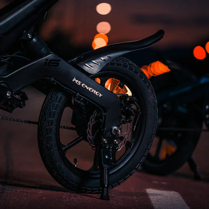 Na sliki je električno kolo znamke MS Energy, model Urbanfold i6. Prikazana je zadnja kolesa in del okvirja, ki je črn in eleganten. Vidni so tudi elementi, kot so zavore in pnevmatika, kar nakazuje na robustno zasnovo, primerno za urbano vožnjo. Osvetlitev v ozadju dodaja dinamičen občutek nočne vožnje.