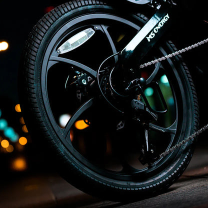 Izdelek na sliki je električno kolo, model MS Energy Urbanfold i6. Na sliki je vidna zadnja kolesa s črnimi platišči in pnevmatikami, ter del prenosa in zavore. Osvetlitev in mestna okolica v ozadju dodajata dinamičen občutek nočne vožnje.