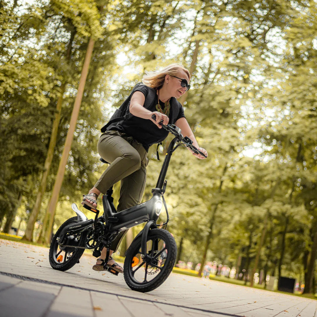 Izdelek na sliki je električno kolo, model MS Energy Urbanfold i6. Na sliki vidimo žensko osebo, ki vozi to električno kolo po urejeni poti v parku. Kolo je zložljivo, ima kompaktno zasnovo in je opremljeno z lučjo spredaj, kar je vidno na sliki. Ozadje je zeleno in naravno, kar ustvarja prijetno vzdušje za vožnjo.