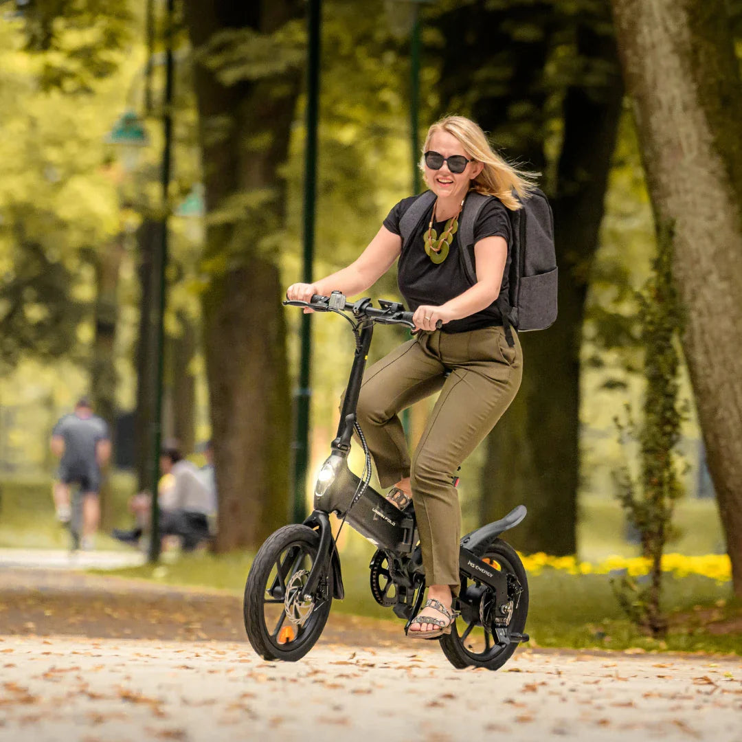 Izdelek je električno kolo, znamke MS Energy, model Urbanfold i6. Na sliki je vidna ženska oseba, ki se vozi na kolesu po urejeni poti v parku, obdana z zelenjem. Oblečena je v udobna oblačila in nosi nahrbtnik, kar nakazuje na sproščeno in aktivno preživljanje časa na prostem. Kolo ima manjše kolesa in kompakten dizajn, kar je primerno za mestno vožnjo.