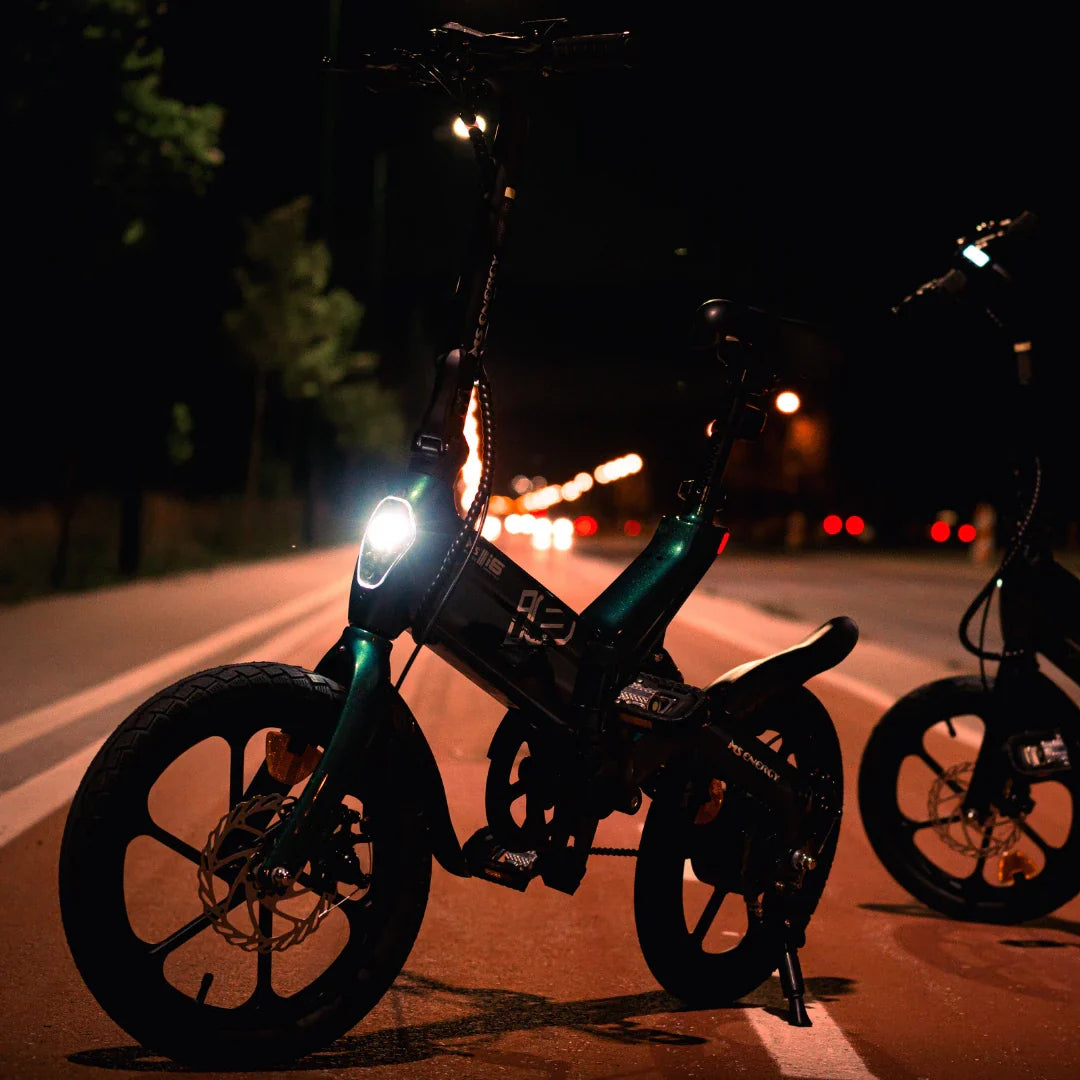 Na sliki je električno kolo, model MS Energy Urbanfold i6. Izdelek je vidno zasnovan za urbano vožnjo, kar se kaže v njegovem kompaktnem dizajnu in zložljivih lastnostih. Kolo ima LED luč spredaj, kar omogoča vožnjo tudi ponoči. Ozadje prikazuje cesto z uličnimi lučmi, kar dodatno poudarja urbano okolje.