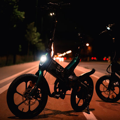 Na sliki je električno kolo, model MS Energy Urbanfold i6. Izdelek je vidno zasnovan za urbano vožnjo, kar se kaže v njegovem kompaktnem dizajnu in zložljivih lastnostih. Kolo ima LED luč spredaj, kar omogoča vožnjo tudi ponoči. Ozadje prikazuje cesto z uličnimi lučmi, kar dodatno poudarja urbano okolje.