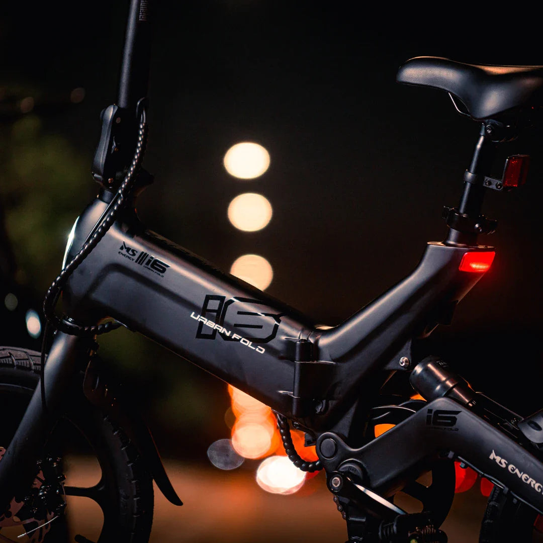 Izdelek na sliki je električno kolo, znamke MS Energy, model UrbanFold i6. Na sliki je vidna struktura okvirja kolesa, ki ima črno barvo in je zasnovano za urbano uporabo. Opremljeno je z zložljivo funkcijo, kar omogoča enostavno shranjevanje in prenašanje. V ozadju so mehke svetlobe, kar ustvarja nočno vzdušje.