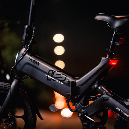 Izdelek na sliki je električno kolo, znamke MS Energy, model UrbanFold i6. Na sliki je vidna struktura okvirja kolesa, ki ima črno barvo in je zasnovano za urbano uporabo. Opremljeno je z zložljivo funkcijo, kar omogoča enostavno shranjevanje in prenašanje. V ozadju so mehke svetlobe, kar ustvarja nočno vzdušje.